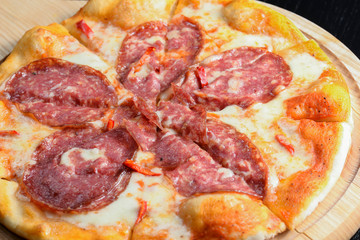 Sliced whole salami pizza on a rustic wooden board over black wooden table. Fast food, junk food concept.