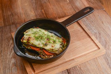 Backed eggplant with mozzarella cheese, tomatoes and basil served in a pan over rustic wooden board in a restaurant.