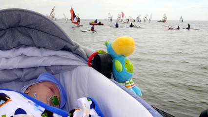 Dziecko w wózku śpi na plaży z widokiem na morze i ludzi uprawiających windserfing.