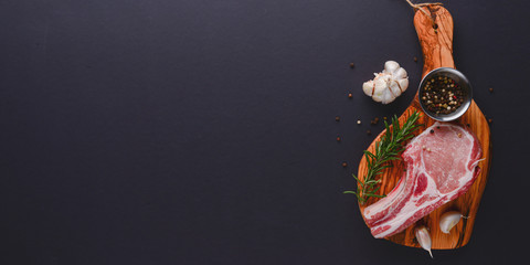 Fresh raw beef steak on wooden cutting board over black background with spices, top view.