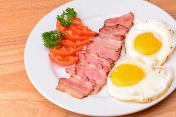 Breakfast with bacon, fried egg and tomatoes on served on white plate over light rustic wooden table.