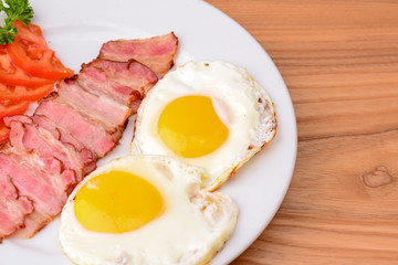 Breakfast with bacon, fried egg and tomatoes on served on white plate over light rustic wooden table.