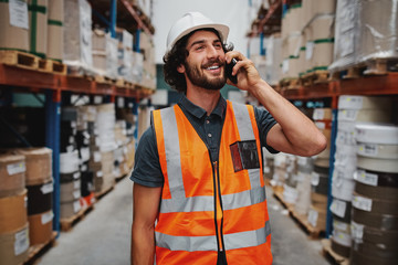 Smiling warehouse manager in conversation on mobile phone wearing white helmet and safety vest standing in between goods shelf looking away