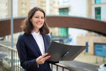 Cheerful businesswoman holding folder. Smiling middle aged businesswoman in formal wear holding papers and looking aside on street. Professional occupation concept