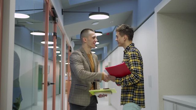 Young man walking down office corridor, browsing through notepad and meeting colleague. Two men greeting each other with handshake, talking cheerfully and saying goodbye