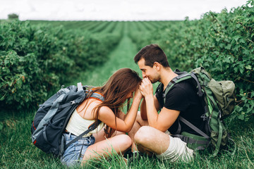 Man and woman with backpacks on their backs sitting hugging on the green grass between the currant bushes. Walking on the currant plantations in the countryside