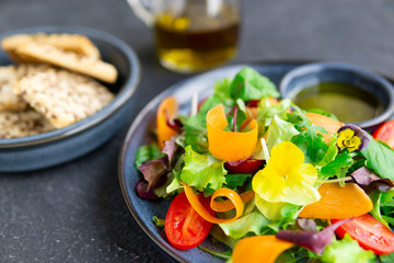 Fresh vegetable salad with tomatoes, carrot slices, greens, flowers, olive oil with herbs, balsamic cream, blue plate, dark textured table. Healthy, diet, vegan, organic food concept. Close up view.