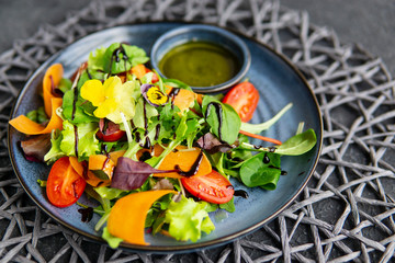 Fresh vegetable salad with tomatoes, carrot slices, greens, flowers, olive oil with herbs, balsamic cream, blue plate, dark textured table. Healthy, diet, vegan, organic food concept. Close up view.