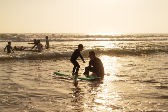 Father looking at sun standing on surfboard. Happy father teaching cute little son in wetsuit surfing on sea waves during sunset. Surfing concept - Powered by Adobe