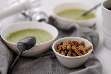 Spring detox broccoli green cream soup in white plates, bowl with crackers and stylish black cutlery on grey background. 