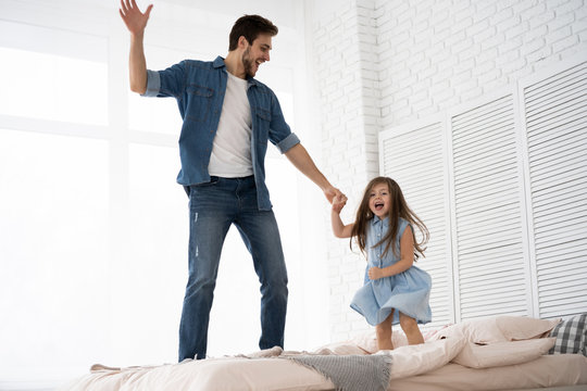 I Love You, Dad! Handsome Young Man At Home With His Little Girl Are Having Fun And Jumping On Bed. Happy Father's Day!