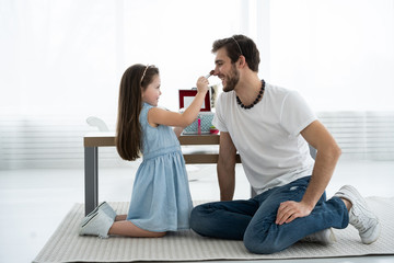 Cute little daughter and her handsome young dad in crowns are playing together in child's room. Girl is doing her dad a makeup.