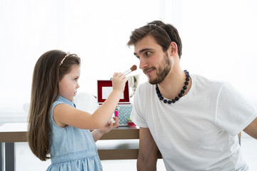 Cute little daughter and her handsome young dad in crowns are playing together in child's room. Girl is doing her dad a makeup.