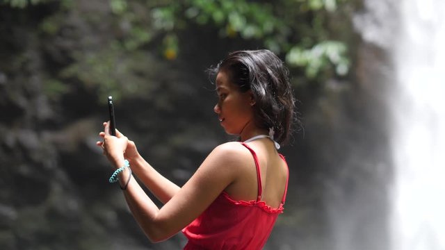 Half body shot at a girl in a waterfall taking selfie photos using smartphone.