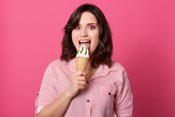 Portrait of delighted attractive hungry young woman standing isolated over pink background in studio, looking directly at camera, licking ice cream, wearing striped shirt. People and food concept.