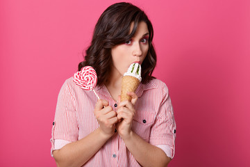 Image of hungry funny beautiful female standing isolated over pink background in studio, holding lollipop and ice cream, eating ice cream, having sweet tooth, wearing striped shirt. Food concept.