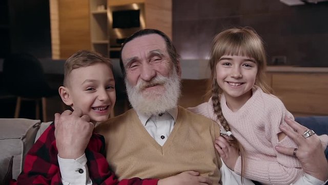 Front view of attractive respected retired bearded man which posing with his two smiling happy good-looking grandchildren on the room background