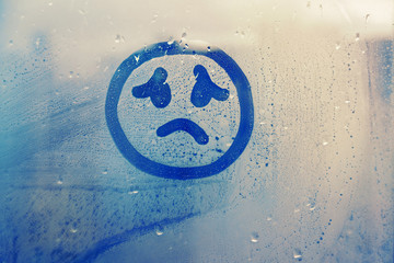 drawn sad face on foggy glass window raindrops blue color concept photo