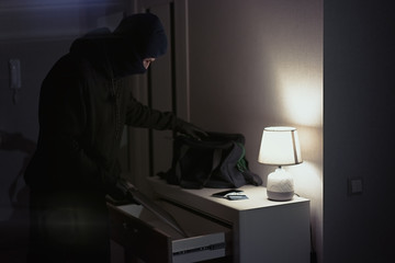 Burglar stealing and putting stolen laptop in bag  inside the house at night - obrazy, fototapety, plakaty