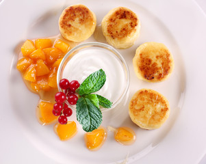 Cheese cakes with sour cream, slices of sweet peach, mint and red currants on a white plate.