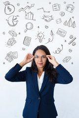 Woman putting on glasses with hand drawn business sketches. Positive confident beautiful young Latin businesswoman in office jacket adjusting eyeglasses with both hands. Glasses wearing concept