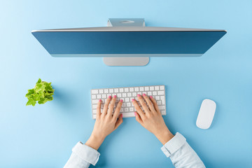 Overhead shot of woman’s hands working on computer with business accessories on blue background....