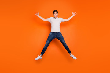 Fototapeta na wymiar Full length photo of crazy funky guy jump high up spread hands legs good mood open mouth wear striped t-shirt jeans sneakers isolated bright orange color background