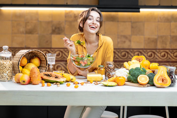 Portrait of a young cheerful woman eating salad at the table full of healthy raw vegetables and...