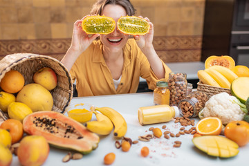 Portrait of a joyful woman with variety of exotic fruits and vegetables on the kitchen, holding sliced cucumis metuliferus. Concept of vegetarianism and healthy eating