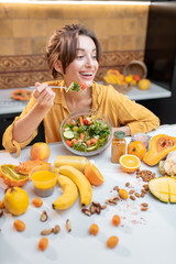 Portrait of a young cheerful woman eating salad at the table full of healthy raw vegetables and fruits on the kitchen at home. Concept of vegetarianism, healthy eating and wellness