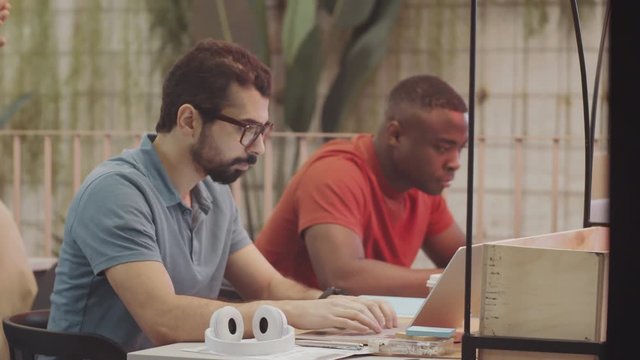 Young bearded middle eastern man in glasses typing on laptop at desk in coworking space while African American colleague working beside him