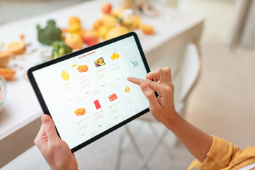 Woman shopping food online using a digital tablet at the kitchen, close-up view on a tablet screen. Concept of buying online using mobile devices