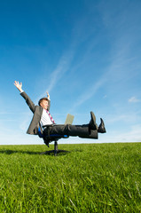 Happy businessman celebrating online success sitting on an office chair outdoors with his laptop computer in the middle of a green meadow