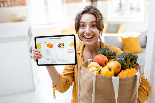 Happy woman holding a digital tablet with launched online store while standing with shopping bag full of fresh products at home. Concept of buying online using mobile devices