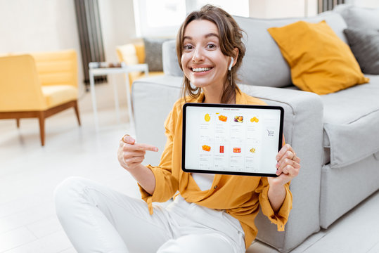 Young And Cheerful Woman Showing A Digital Tablet Screen With Launched Online Store, Shopping Online At Home. Concept Of Buying Online Using Mobile Devices