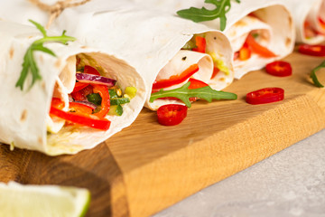 Fresh vegan tortilla wraps with bell pepper, tomato and cucumber on wooden cutting board with lime and chili pepper. Light grey stone background. Close up.