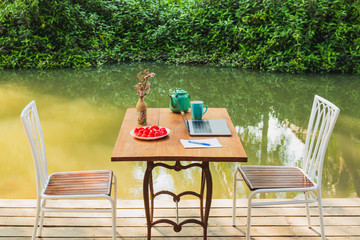 Laptop computer with tea mug and rose apple fruit on wood table by the canal.
