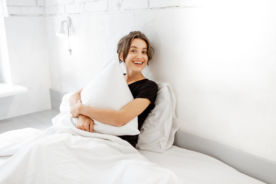Portrait of a young and cute woman hugging a pillow while sitting relaxed in bed at the white bedroom