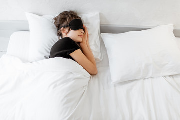 Woman lying with a sleeping mask in bed covered with white sheets in bright bedroom in the morning