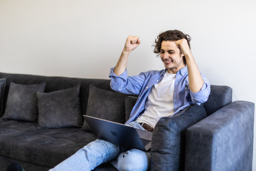 Happy excited young man win gesture celebrate with laptop at home sitting on a couch