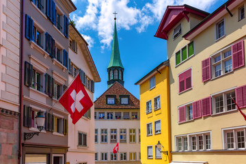 Swiss flag waving, church and colorful facades of houses in Old Town of Zurich, the largest city in...