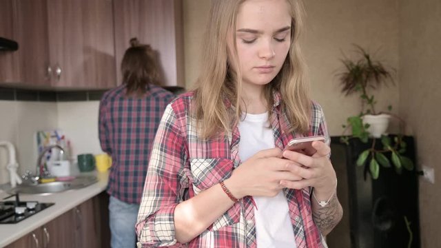 Young married couple in the kitchen. A girl with a tattoo writes a message on her mobile phone to her lover on the background of her husband who washes the dishes.