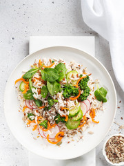 Appetizing healthy salad of white and wild rice with vegetables, green pesto sauce and seeds in white plate on a light background. Selective focus. Top view.