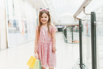 Little fashion girl with packages in a large shopping center. Pretty smiling little girl with shopping bags posing in the shop. child in pink dress near shopping mall having fun