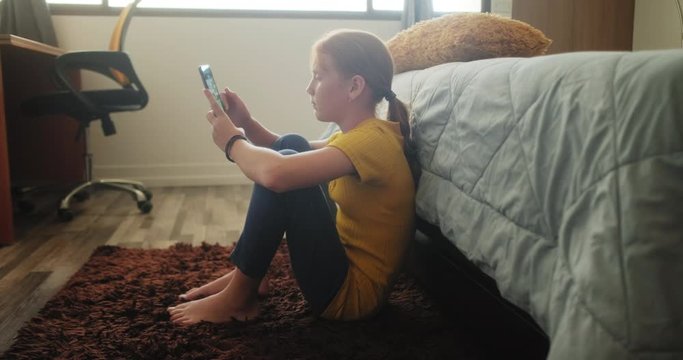 Text message on mobile telephone for bullying a sad little girl in bedroom at home. Young people looking at cell phone. Redhead female child with emotions, feelings of sadness and loneliness