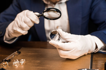 Cropped view of jewelry appraiser holding gemstone and magnifying glass near jewelry rings on board...