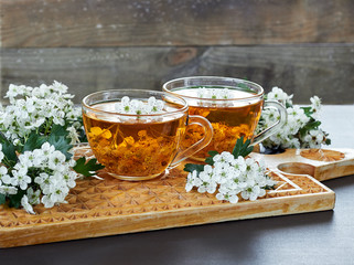 Fototapeta premium Two cups of herbal haw thorn tea from flower buds around blooming branches on wooden cutting board on rustic background, closeup, copy space, heart health and healthy herbal drinks concept