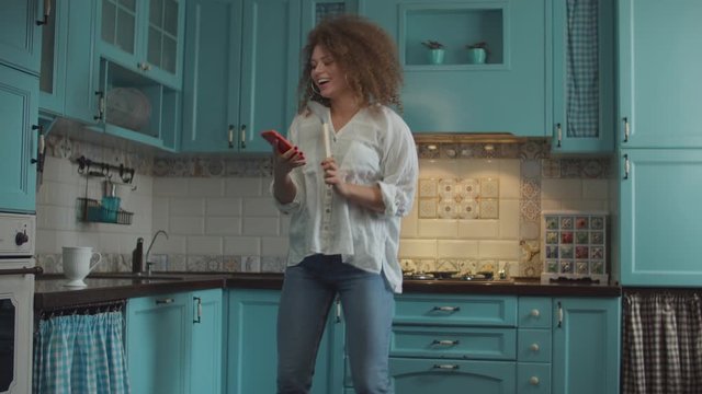 Smiling curly 20s woman in jeans dancing happily with scoop as microphone and mobile phone in hands on home blue kitchen. 