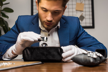 Handsome jewelry appraiser holding magnifying glass while examining gemstone near earrings on table