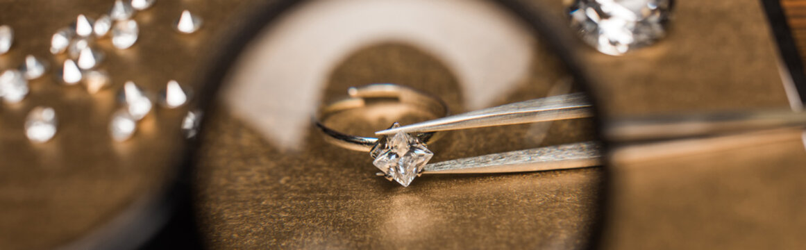 Selective focus of magnifying glass, jewelry ring with gemstone in tweezers on board, panoramic shot